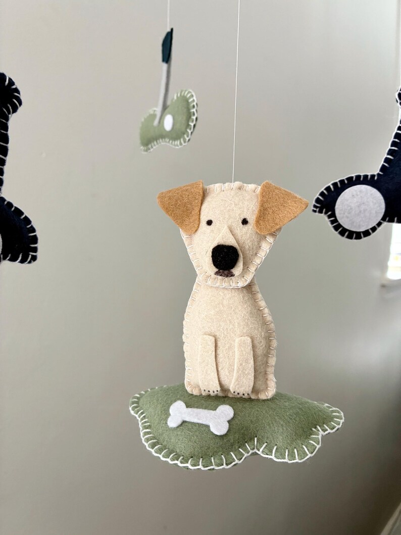 May include: A handmade felt dog mobile featuring a cream-colored dog with tan ears and a black nose. The dog sits on a green cloud with a white bone. Other felt shapes, including a bone and a dark shape, hang above.