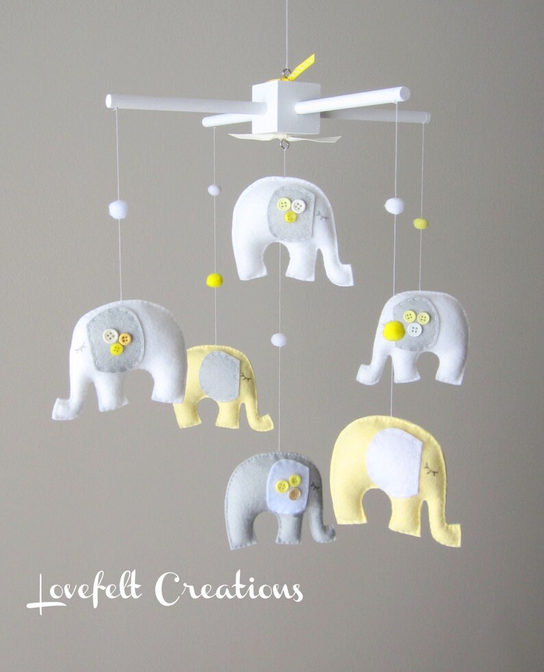 May include: A white wooden baby mobile with six gray and yellow felt elephant ornaments. The elephants have button eyes and are suspended from the mobile by white strings.