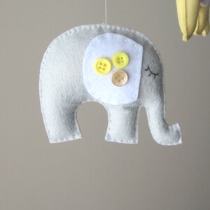 May include: A grey felt elephant with white felt ears and a white felt patch on its back. The patch has three yellow buttons and one brown button.