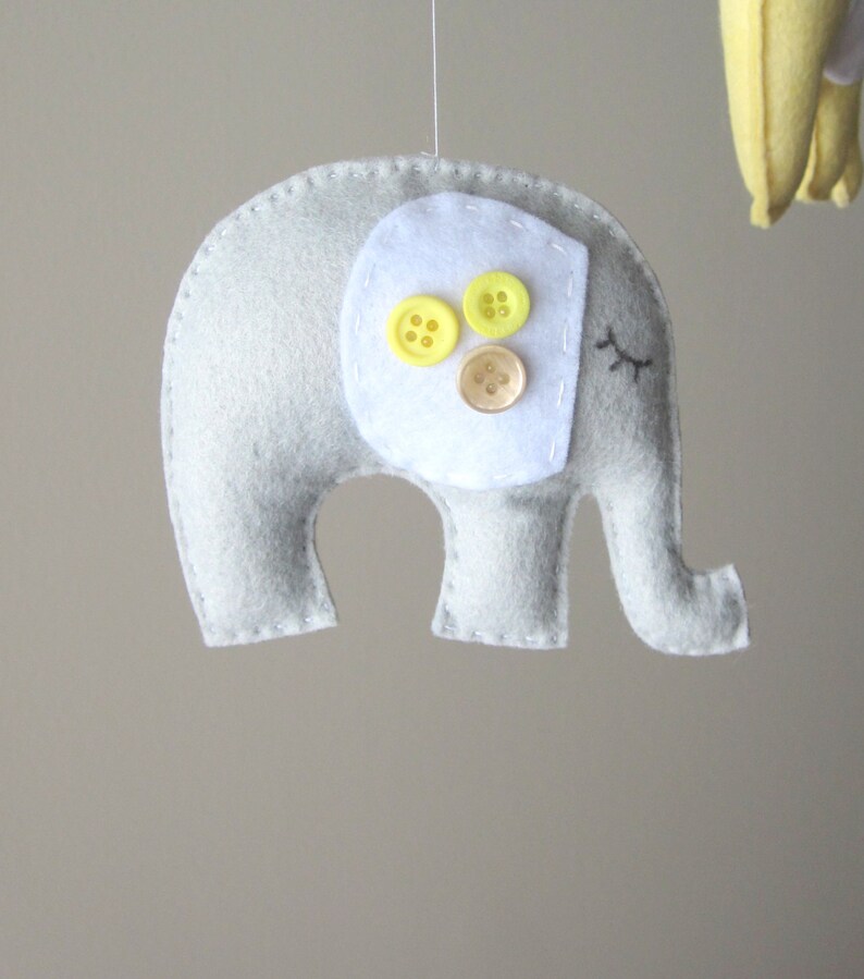 May include: A grey felt elephant with white felt ears and a white felt patch on its back. The patch has three yellow buttons and one brown button.
