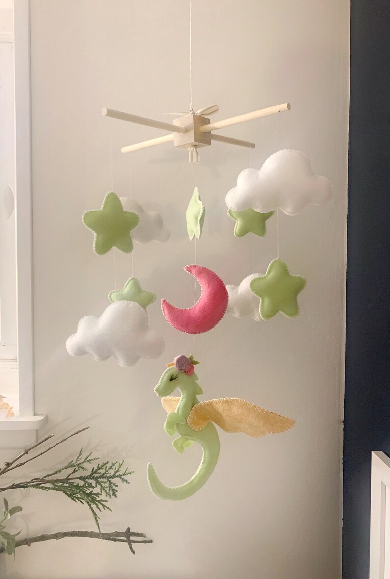May include: A green felt dragon with yellow wings and a pink felt moon hangs from a wooden mobile with white felt clouds and green felt stars.