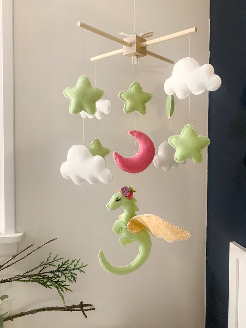 May include: A green felt dragon with yellow wings and pink flowers on its head hangs from a wooden mobile with white clouds, green stars, and a pink crescent moon.