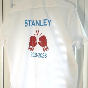 Könnte beinhalten: Weißes kurzärmeliges Hemd mit dem Namen "STANLEY" in blauer Schrift und einem roten Boxhandschuh-Motiv. Die Jahre "2021-2026" sind darunter aufgedruckt. Das Hemd hängt auf einer weißen Oberfläche.