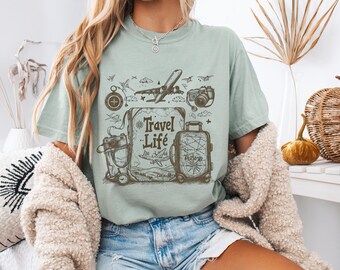 Könnte beinhalten: Hellgrünes T-Shirt mit einem Reise-Themen-Motiv. Die Grafik zeigt einen Kompass, ein Flugzeug, eine Kamera, einen Koffer und die Worte "Travel Life". Das Shirt hat einen lässigen Stil.