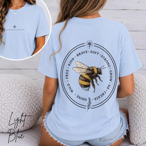 May include: A light blue t-shirt featuring a small flower graphic and the text "BLOOM WHERE YOU ARE PLANTED" on the front. The back displays a large circular design with a central bee illustration surrounded by the words "KIND", "BRAVE", "SOFT", "STRONG", "GROW", "CREATE", "SHINE", "WILD", and "FREE".