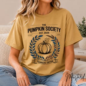 May include: A mustard yellow t-shirt featuring a graphic with the text THE PUMPKIN SOCIETY EST. 2026 and FOUNDERS EDITION. The design includes a central pumpkin illustration surrounded by a circular wreath of blue and brown leaves and wheat.