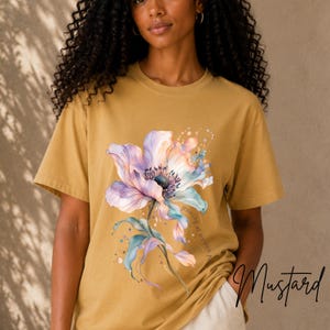Puede incluir: Una camiseta color amarillo mostaza con una ilustración estilo acuarela de una flor grande de color azul y violeta claro con tallos verdes. El texto "growing at my own pace" está impreso en una fuente cursiva pequeña junto al gráfico.