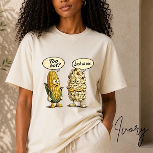 Op de afbeelding: Een ivoorkleurig t-shirt met een afbeelding van een gele maïskolf met een tekstballon "Too hot?" naast een stuk popcorn met een tekstballon "Look at me.". De maïs kijkt verbaasd, terwijl de popcorn er geërgerd uitziet.