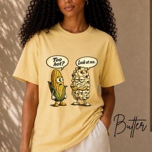 May include: A light yellow t-shirt featuring a graphic of a cartoon corn cob with the text "Too hot?" and a cartoon piece of popcorn with the text "Look at me." The corn cob has a surprised expression, while the popcorn appears annoyed.