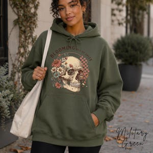 May include: An olive green hooded sweatshirt featuring a graphic print of a human skull surrounded by colorful flowers and a checkered pattern. The text "BLOOMING OUT OF SPITE" is arched above the skull, and a plain white tote bag is on the shoulder.