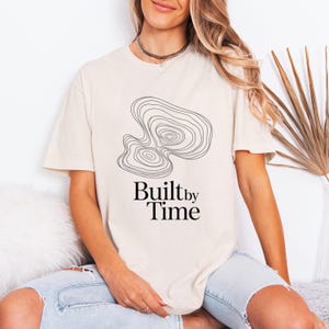 以下が含まれることがあります： 黒い線画のデザインと「Built by Time」の文字が入ったクリーム色のTシャツ。グラフィックは地形図に似ています。柔らかい素材でできており、ゆったりとしたフィット感です。