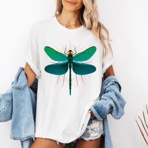 May include: White t-shirt featuring a teal and green dragonfly graphic. The dragonfly has detailed wings and a geometric design. The shirt is a casual style, with rolled-up sleeves. The dragonfly design is centered on the front of the shirt.