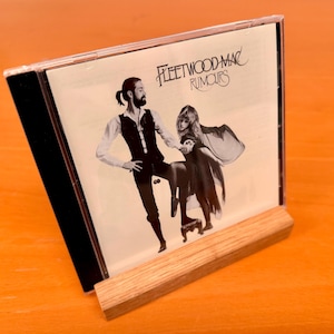 Puede incluir: Un estuche de CD del álbum "Rumours" de Fleetwood Mac exhibido en un soporte de madera. La portada del álbum presenta una imagen en blanco y negro de dos personas. El soporte es de madera clara.