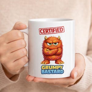 Peut inclure: Mug en céramique blanche avec un dessin de monstre de dessin animé. Le monstre a des poils orange, des cornes jaunes et les bras croisés. Le mug porte l'inscription "CERTIFIED" dans un tampon rouge et "GRUMPY BASTARD" en jaune et bleu.