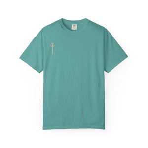 Puede incluir: Una camiseta turquesa de manga corta con un pequeño gráfico de tridente de color claro en el pecho izquierdo. La camiseta tiene cuello redondo y un corte relajado. La tela parece ser un material suave y cómodo.