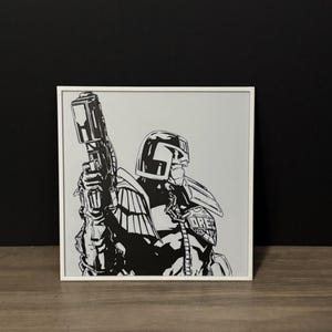 May include: Black and white framed print of a comic book character holding a large gun. The character is wearing a helmet and uniform. The artwork is displayed in a white frame.