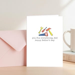 Könnte beinhalten: Eine weiße Vatertagskarte mit einer farbenfrohen Illustration von Werkzeugen, darunter ein Schraubenschlüssel, ein Schraubendreher, ein Hammer und eine Säge. Auf der Karte steht "you fix everything, dad happy father's day". Ein rosa Umschlag und eine Tasse sind im Hintergrund.