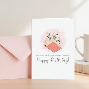 Pode incluir: Um cartão de felicitações branco com o texto "Happy Birthday!" e "Another exciting chapter begins..." em uma fonte de script. O cartão apresenta uma ilustração de um livro aberto com flores e um fundo rosa em aquarela. Um envelope rosa está à esquerda.