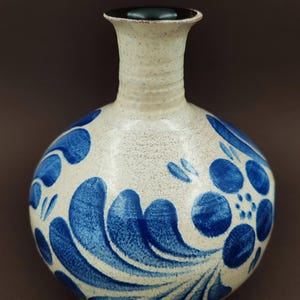 Könnte beinhalten: Eine Keramikvase mit rundem Körper und schmalem Hals. Die Vase ist cremefarben mit gesprenkelter Textur und mit blauen, stilisierten Blumenmustern verziert. Der Rand ist dunkelbraun.