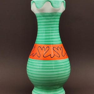 Può includere: Un vaso in ceramica con corpo a righe verdi e bianche, una fascia arancione con disegni floreali neri e un bordo bianco smerlato. Il vaso ha una base bulbosa e un piede svasato.