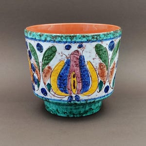 Könnte beinhalten: Ein Keramik-Blumentopf mit einem lebendigen Blumendesign. Der Topf hat einen weißen Hintergrund mit bunten Blumen in Gelb, Blau und Rosa, akzentuiert durch grüne Blätter und blaue Punkte. Der Rand ist orange und der Sockel grün.