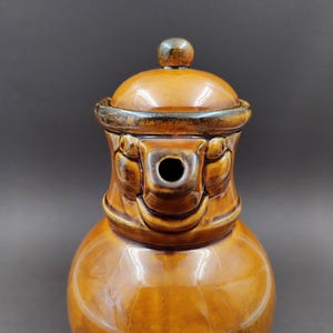Könnte beinhalten: Eine braune Keramik-Teekanne mit Deckel und Ausguss. Die Teekanne hat einen abgerundeten Körper und ein dekoratives Design um den oberen Rand. Der Deckel hat einen kleinen, runden Knopf. Die Teekanne ist glasiert mit einem glänzenden Finish.