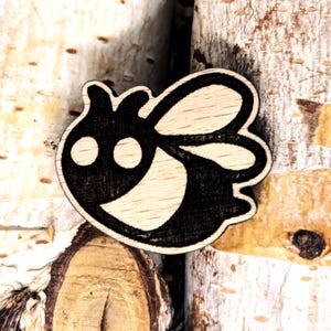 Puede incluir: Un broche de madera con forma de abeja con contorno y detalles negros. La abeja tiene dos ojos grandes y una boca sonriente. Las alas también están delineadas en negro. El broche está hecho de madera clara.