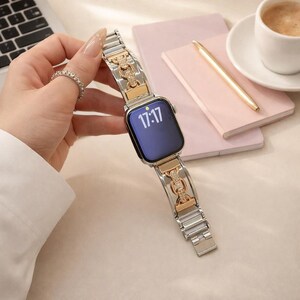 May include: Silver and gold-toned watch band with a decorative chain link design. The watch face displays the time as 17:17. The band is held by a hand wearing a silver ring.