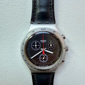 May include: A Swatch chronograph watch with a black dial, silver case, and black leather strap. The watch face features multiple sub-dials and a red second hand. The text "Swatch Swiss" is visible on the dial.