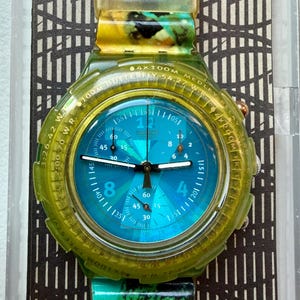 Puede incluir: Un reloj Swatch con esfera azul y caja y correa verde transparente. La esfera del reloj presenta números y marcas, con el logotipo de Swatch en la parte superior. La correa tiene un patrón verde y amarillo veteado.