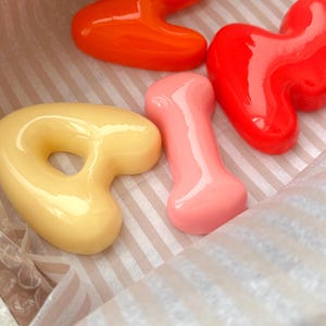Op de afbeelding: Een assortiment kleurrijke, glanzende, abstract gevormde zepen in een gestreepte doos. De zepen zijn in tinten oranje, rood, geel en roze. De vormen omvatten een acht, een hart, een bot en een donut.