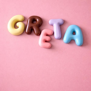 Puede incluir: El nombre "GRETA" escrito con letras de burbujas 3D de colores. Las letras son amarillas, marrones, rosas, moradas y azules, sobre un fondo rosa liso. Las letras son redondeadas y tienen un acabado brillante.
