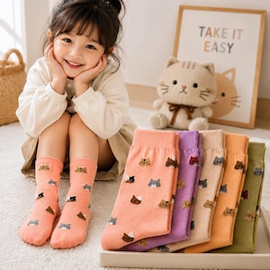 Peut inclure: Un ensemble de chaussettes montantes avec un motif répétitif de petites têtes de chat dans diverses couleurs, notamment pêche, violet, beige, orange et vert. Les chaussettes sont en tricot doux et présentées en pile pliée avec une paire pêche.