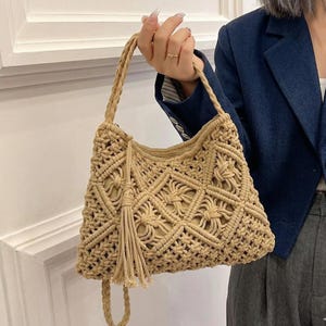 May include: A beige macrame handbag with a woven design and a braided handle. The bag features a tassel and a rectangular shape. The bag is held by a person wearing a navy blue blazer.