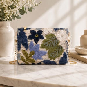 Pode incluir: Uma bolsa clutch retangular com uma base tecida em tom creme e bordados florais intrincados em tons de azul-marinho, azul-claro e verde-oliva. O design inclui detalhes em lantejoulas, um fecho de correr dourado e uma alça de corrente dourada.