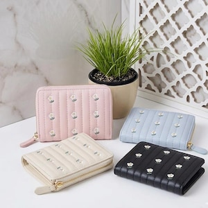 Op de afbeelding: Vier kleine portemonnees met ritssluiting in pastelkleuren: roze, lichtblauw, crème en zwart. Elke portemonnee heeft een gewatteerd ontwerp met geborduurde witte bloemaccenten. De portemonnees liggen op een wit oppervlak.