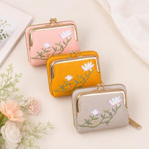 May include: Three small rectangular wallets with gold-tone metal kiss-lock closures. The wallets are pink, mustard yellow, and light gray, each featuring a textured surface and embroidered white flowers with green stems and leaves on the front.