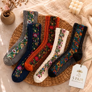 Op de afbeelding: Vijf paar gebreide sokken in grijs, marineblauw, oranje, crème en zwart, elk met een verticaal paneel van kleurrijk bloemenborduursel. Een wit rechthoekig label is bevestigd met de tekst 5 PAIR DELUXE COLLECTION en een klein icoontje.