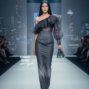 May include: A model in a gray, one-shoulder gown with a sheer, ruffled sleeve and a matching handbag. The gown has a fitted bodice and a long, straight skirt. The text "SIHAM AYAD" is visible.
