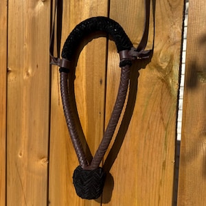 May include: A black and brown horse halter with a braided leather design. The halter features a black padded crown and noseband, with brown leather straps and accents. The halter is hanging against a wooden background.