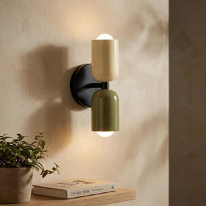 Könnte beinhalten: Moderne Wandleuchte mit schwarzer Montageplatte. Die Leuchte hat zwei zylindrische Leuchten, eine cremefarbene und eine olivgrüne, jeweils mit einer runden Glühbirne. Die Leuchte ist an einer beigefarbenen Wand montiert.