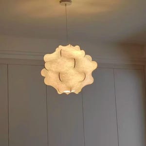 Op de afbeelding: Een hanglamp met een uniek, wolkenachtig ontwerp. De crèmekleurige, getextureerde kap heeft een geschulpte rand. De lamp hangt aan een dun, wit snoer en straalt een warme gloed uit.