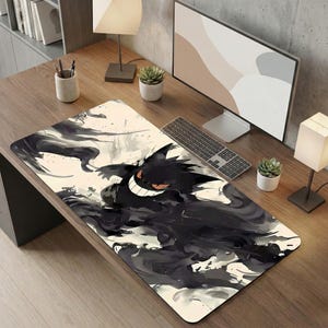 Puede incluir: Alfombrilla de ratón rectangular con una ilustración en blanco y negro de Gengar, mostrando una amplia sonrisa. El diseño tiene un estilo acuarela con patrones oscuros y arremolinados. La alfombrilla está sobre un escritorio de madera con un ordenador y teclado.
