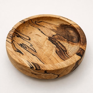 May include: A round wooden bowl featuring a light tan base color with prominent dark brown and black spalted grain patterns throughout the surface. The bowl has a smooth, shallow interior basin and a thick, rounded rim with a natural wood texture.