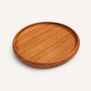 May include: A round wooden tray with a smooth, polished surface and a visible natural grain pattern. The tray features a shallow, concave center and a raised, rounded rim, showcasing a warm, medium-brown tone throughout the entire object.