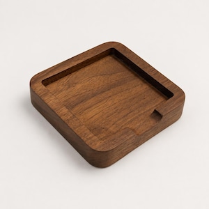 May include: A square wooden coaster crafted from dark brown walnut with a visible natural wood grain. The coaster features a recessed center area with rounded corners and a small notch cut into one side of the raised outer rim edge.