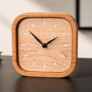 Op de afbeelding: Een vierkante houten klok met een lichtbruine houtnerf wijzerplaat en een bijpassende lijst. De wijzerplaat heeft witte markeringen en zwarte wijzers. De klok staat op een donkere ondergrond.
