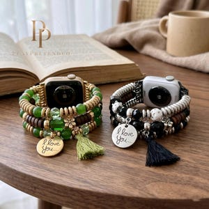May include: Two Apple Watch bands with beaded designs. One band features green beads, gold accents, and a green tassel. The other band has black and silver beads, brown accents, and a black tassel. Both bands have a charm that says "I love you..."