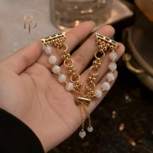 May include: A gold-toned watch band featuring two strands of white cat's eye beads and a central strand of gold-toned metal rings and small spheres. The band includes an adjustable sliding clasp with two dangling chains tipped with white beads.