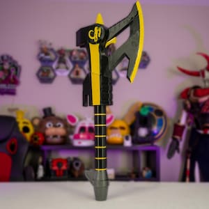 May include: A large, prop axe with a black and yellow color scheme. The axe head is black with yellow accents, and the handle is black with yellow stripes. The axe has a gray base and a yellow emblem. The background is blurred, with various character masks and figures.